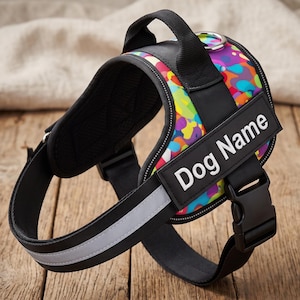 May include: A dog harness featuring a vibrant multicoloured camouflage pattern with black trim and straps. It includes a top handle, a silver metal D-ring, a reflective strip along the bottom, and a black rectangular patch with the text Dog Name.