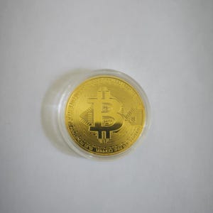 Può includere: Una moneta Bitcoin dorata in una custodia di plastica trasparente.