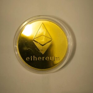 Può includere: Una moneta dorata con il logo di Ethereum e la parola "ethereum" incisa su di essa. La moneta è in una custodia di plastica trasparente.