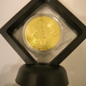 May include: A gold-colored Bitcoin coin is displayed in a black diamond-shaped display case with a black base. The coin has the Bitcoin symbol on it.