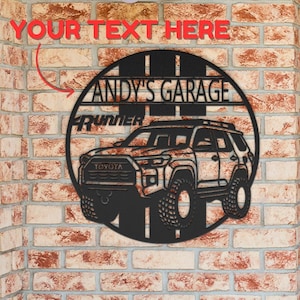 May include: A circular black metal wall sign featuring a cutout design of a Toyota 4Runner SUV. The sign includes the text "ANDY'S GARAGE" and "4RUNNER" integrated into the design. The metal art piece is mounted on a textured brick wall surface.
