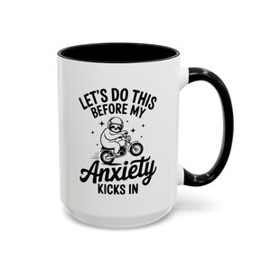 May include: White coffee mug with a black interior, handle, and rim. The mug features the text "LET'S DO THIS BEFORE MY ANXIETY KICKS IN" with a sloth riding a bicycle graphic. A fun and quirky design.