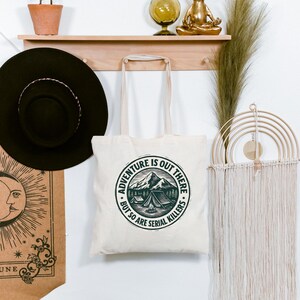 May include: A cream-colored tote bag with the text "Adventure is out there, but so are serial killers" in a circular design featuring mountains and a tent. The bag has two handles and is hanging on a white wall.