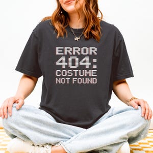 May include: Dark grey t-shirt featuring the text "ERROR 404: COSTUME NOT FOUND" in a pixelated white font. The shirt has short sleeves. The person is wearing light blue jeans.