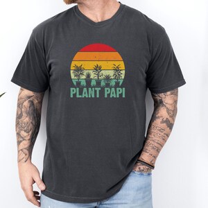 May include: Dark gray t-shirt with a retro sunset design featuring potted plants and the text "PLANT PAPI" in green. The shirt has a v-neck and short sleeves. The design is centered on the chest.