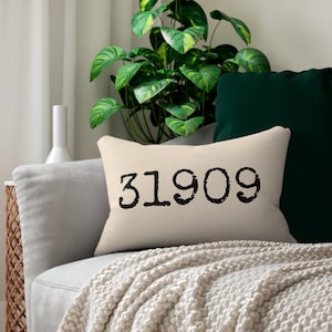 Puede incluir: Un cojín rectangular beige con el número "31909" impreso en negro. El cojín está sobre un sofá gris, con una planta verde y un cojín verde oscuro en el fondo. También se ve un jarrón blanco.