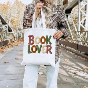 Könnte beinhalten: Eine cremefarbene Tragetasche mit dem Schriftzug "BOOK LOVER" in farbigen, strukturierten Buchstaben. Die Buchstaben sind in Braun-, Grün- und Cremetönen gehalten. Die Tasche wird von einer Person gehalten, die ein braunes kariertes Hemd und eine hellblaue Jeans trägt.
