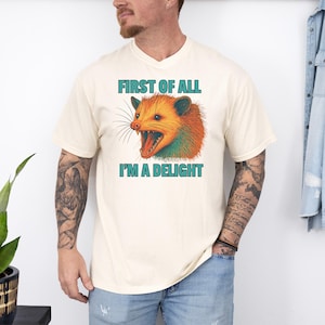 May include: Cream-colored t-shirt featuring a colorful opossum graphic with the text "FIRST OF ALL I'M A DELIGHT" in teal. The opossum has orange, green, and blue fur, with an open mouth. The shirt is a casual, unisex style.