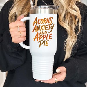 May include: A white travel mug with a handle and a silver lid. The mug features the text "Acorns, Anxiety, and Apple Pie" in orange and brown lettering. The mug is held by a person.