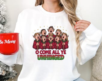 May include: White sweatshirt with a cartoon choir in red robes and the text "O COME ALL YE UNHINGED" in red and green. A red mug with "Be Merry" is held in the left hand. The design is festive and humorous.