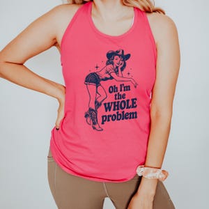 May include: A hot pink racerback tank top with a navy blue graphic of a cowgirl and the text "Oh I'm the WHOLE problem". The cowgirl is wearing a hat, shorts, and boots. The tank top is made of a soft material.