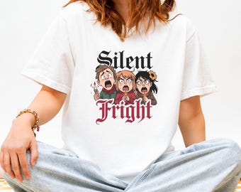 May include: White t-shirt featuring a cartoon graphic with the words "Silent Fright." The graphic depicts three people with shocked expressions, holding candles and ice cream cones. The text is in a gothic font.