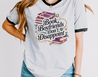 May include: A gray ringer tee with black trim featuring the text "Book Boyfriends Don't Disappoint" in a decorative font. The graphic includes stacks of purple books, keys, and floral accents.