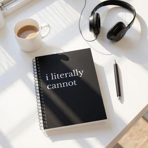 May include: A black spiral-bound notebook with the text "i literally cannot" in white. A white mug with coffee, black headphones, and a black pen are also on the white surface.