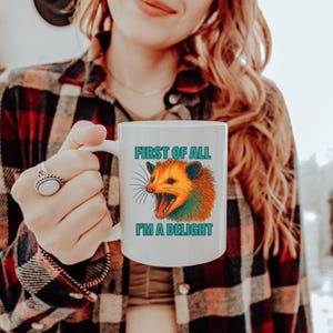 May include: White ceramic mug featuring a colorful opossum illustration with the text "FIRST OF ALL I'M A DELIGHT" in teal. The opossum has orange, green, and brown fur. The mug is held by a person wearing a plaid shirt.