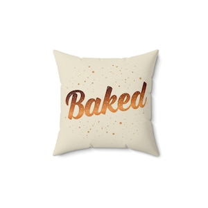 May include: Square throw pillow with the word "Baked" in a gradient brown script. The pillow is cream-colored with small brown speckles scattered across the surface.