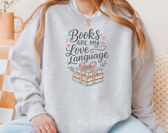 May include: A light gray sweatshirt with the text "Books are my Love Language" in a script font. The design includes a stack of books tied with a pink ribbon and small hearts. The sweatshirt is a casual style.