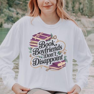 Op de afbeelding: Witte longsleeve met de tekst "Book Boyfriends Don't Disappoint" in een decoratief lettertype. Het ontwerp bevat illustraties van paarse boeken met gouden accenten en sleutels.