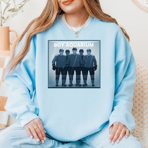 May include: Pale blue sweatshirt with a graphic of five hockey players in blue uniforms, helmets, and black gloves, inside a frame. The text "BOY AQUARIUM" is above the image. The background has a rainy effect.
