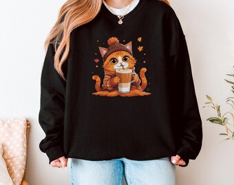 Op de afbeelding: Zwarte sweatshirt met een cartoonkat met oranje vacht, een bruine hoed en een geruite sjaal, die een koffiekopje vasthoudt. Oranje bladeren omringen de kat. De sweatshirt wordt gedragen met een blauwe spijkerbroek.