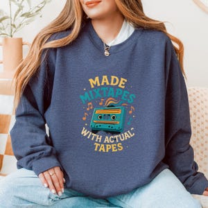 Peut inclure: Sweat-shirt bleu marine avec l'inscription "MADE MIXTAPES WITH ACTUAL TAPES" dans une police rétro. Le motif comprend une cassette turquoise et des notes de musique jaunes et turquoise. Style décontracté et confortable.