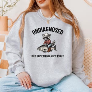 May include: A light gray sweatshirt with the text "UNDIAGNOSED" above an illustration of a raccoon wearing a cowboy hat and riding a fish. The text "BUT SOMETHING AIN'T RIGHT" is below the illustration.