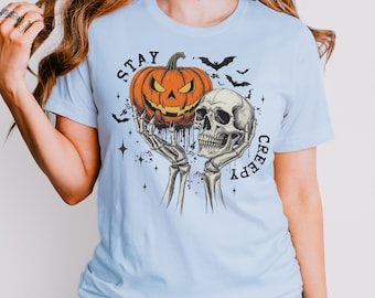 May include: Light blue t-shirt with a Halloween-themed graphic. The design features a carved orange pumpkin, a human skull, bats, and the words "STAY CREEPY". The pumpkin and skull are held by skeletal hands.
