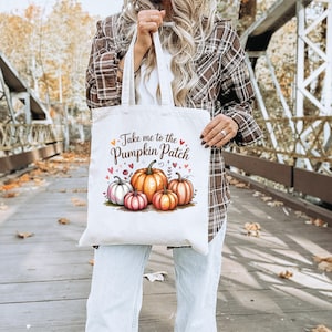 Könnte beinhalten: Eine cremefarbene Tragetasche mit dem Text "Take me to the Pumpkin Patch" und einer Illustration von Kürbissen. Die Kürbisse sind orange, rosa und weiß, mit grünen Blättern und kleinen Herzen.