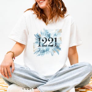 Puede incluir: Camiseta blanca con el número "1221" en negro, centrado en la parte delantera. El número está superpuesto a un diseño abstracto azul y blanco con detalles dorados. El modelo lleva vaqueros azul claro.