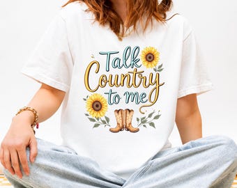 Puede incluir: Una camiseta blanca de manga corta con un estampado gráfico que incluye el texto Talk Country to me en letras azules y doradas. El diseño presenta dos girasoles amarillos con hojas verdes y un par de botas vaqueras marrones en la base.