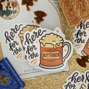 May include: Die-cut vinyl stickers featuring a cartoon mug of frothy orange butterbeer with the text 'here for the' in black script and 'BUTTERBEER' in a decorative frame. The design includes small gold star accents on a white background.