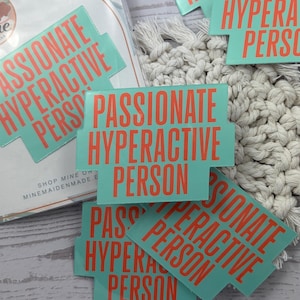 May include: A collection of teal stickers with the words "PASSIONATE HYPERACTIVE PERSON" in orange. The stickers are scattered on a light-colored wooden surface, with a macrame coaster in the background.