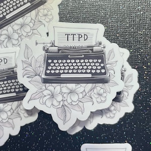 May include: Black and white sticker of a vintage typewriter with the text "TTPD" on the paper in the typewriter. The typewriter is surrounded by stylized flowers.