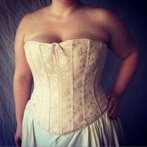 May include: A strapless corset in a pale cream color featuring a subtle, tonal floral jacquard pattern throughout. The garment is constructed with vertical boning channels and includes a small white cord tie at the center of the straight neckline.
