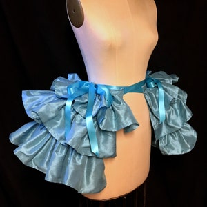 May include: A light blue ruffled skirt with a matching ribbon belt and bows. The skirt is displayed on a mannequin, paired with a black choker necklace with a silver-toned embellishment. The fabric has a slight sheen.