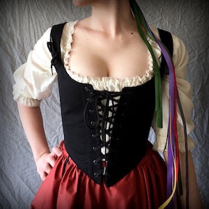 May include: A black corset with a lace-up front and a white lace trim. The corset is worn over a white blouse with a ruffled neckline. The model is wearing a red skirt and colourful ribbons.