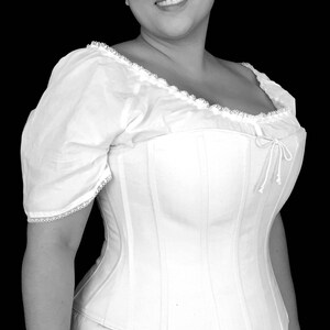 May include: A white cotton corset with a lace trim and a bow at the back. The corset is being worn by a person with dark hair and a white blouse.