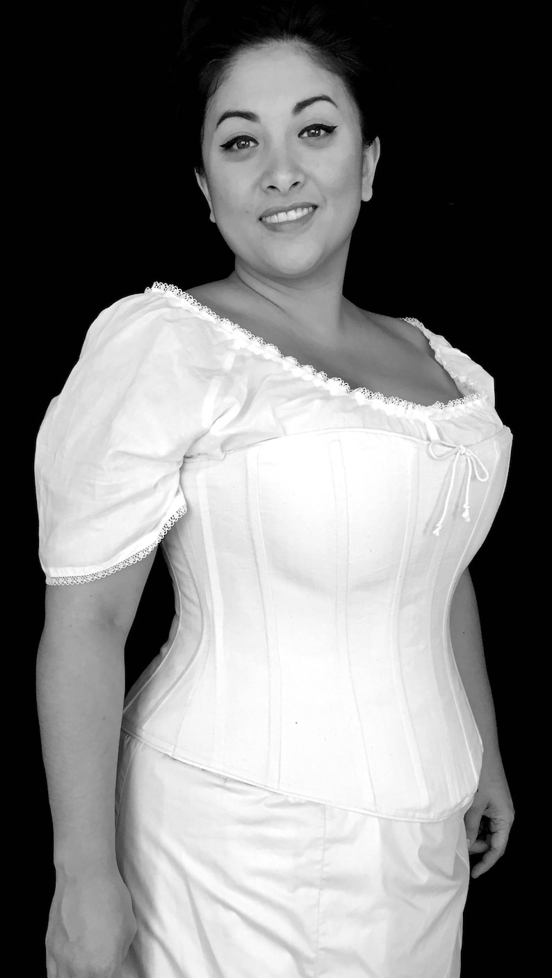 May include: A white cotton corset with a lace trim and a bow at the back. The corset is being worn by a person with dark hair and a white blouse.