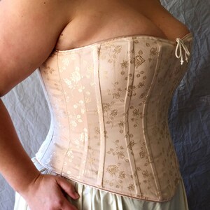 May include: A beige corset featuring a subtle tonal floral pattern with vertical boning channels and a small white cord tie at the center of the neckline. The garment is constructed with structured panels and a straight upper edge design.