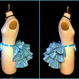 以下が含まれることがあります： 後ろに結ばれたサテンリボン付きの薄いブルーのフリル付きバストル。バストルは黒い背景の白いドレスフォームに着用されています。