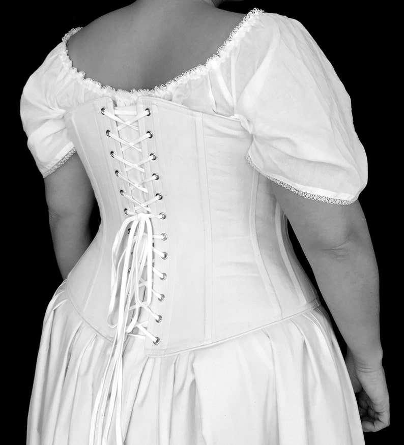 May include: A white corset with a lace-trimmed bodice and a long, flowing skirt. The corset has a lace-up back with white ribbon ties.