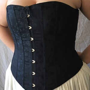 May include: A black strapless corset featuring a subtle tonal floral jacquard pattern throughout the fabric. The garment includes a front-fastening closure with silver-toned metal busk hooks and a small drawstring tie at the top neckline.
