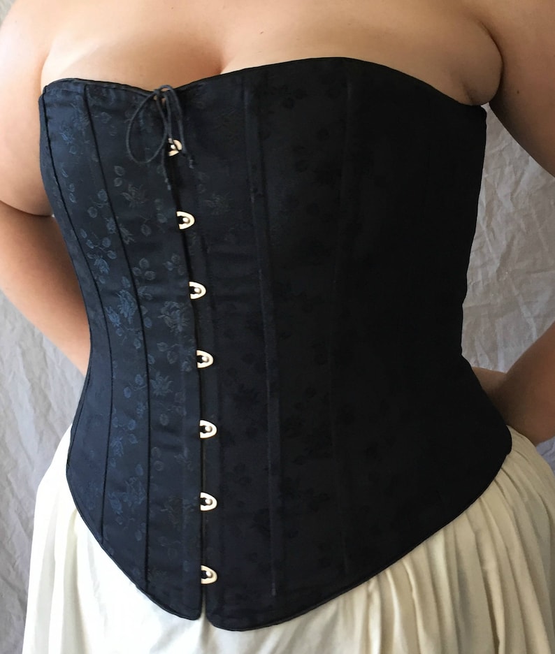 May include: A black strapless corset featuring a subtle tonal floral jacquard pattern throughout the fabric. The garment includes a front-fastening closure with silver-toned metal busk hooks and a small drawstring tie at the top neckline.