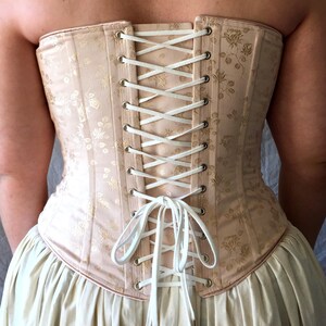 May include: A champagne-colored corset featuring a subtle gold floral pattern. The garment is secured at the back with white lacing threaded through silver-toned metal grommets, tied in a bow at the bottom, and worn over a cream-colored skirt.