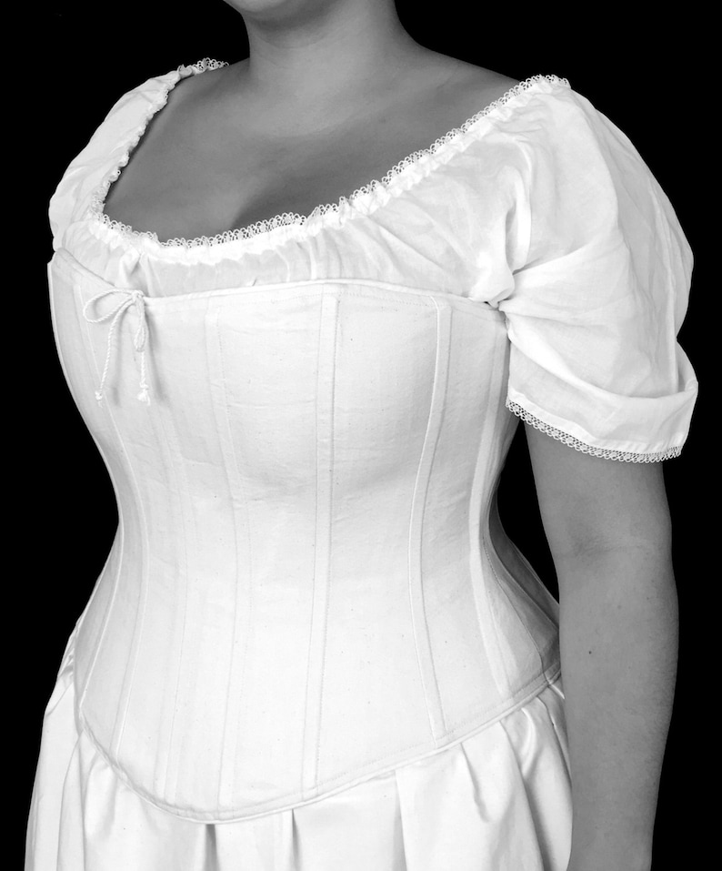 May include: A white cotton corset with a lace trim and a gathered neckline. The corset has a boned structure and a long, flowing skirt.