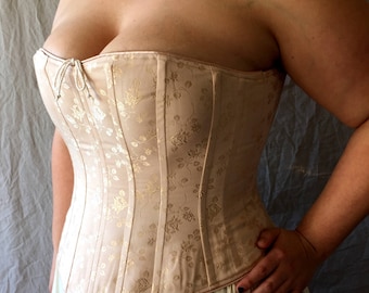May include: A strapless corset in a pale beige hue featuring a subtle, shimmering gold floral jacquard pattern throughout. The garment is constructed with vertical boning channels and includes a small, delicate white cord tie at the center neckline.
