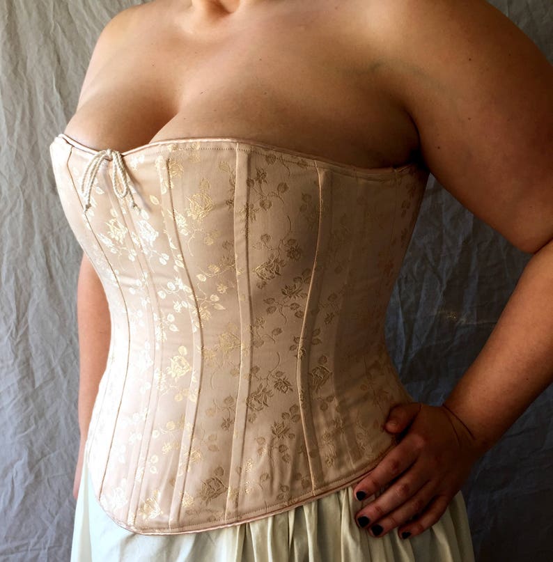 May include: A strapless corset in a pale beige hue featuring a subtle, shimmering gold floral jacquard pattern throughout. The garment is constructed with vertical boning channels and includes a small, delicate white cord tie at the center neckline.