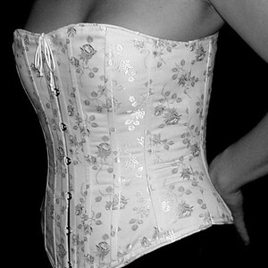 May include: A white corset featuring a tonal floral pattern with a subtle sheen. The garment includes vertical boning channels and a front closure with silver-toned metal busk clasps and a white cord lacing detail at the top neckline.