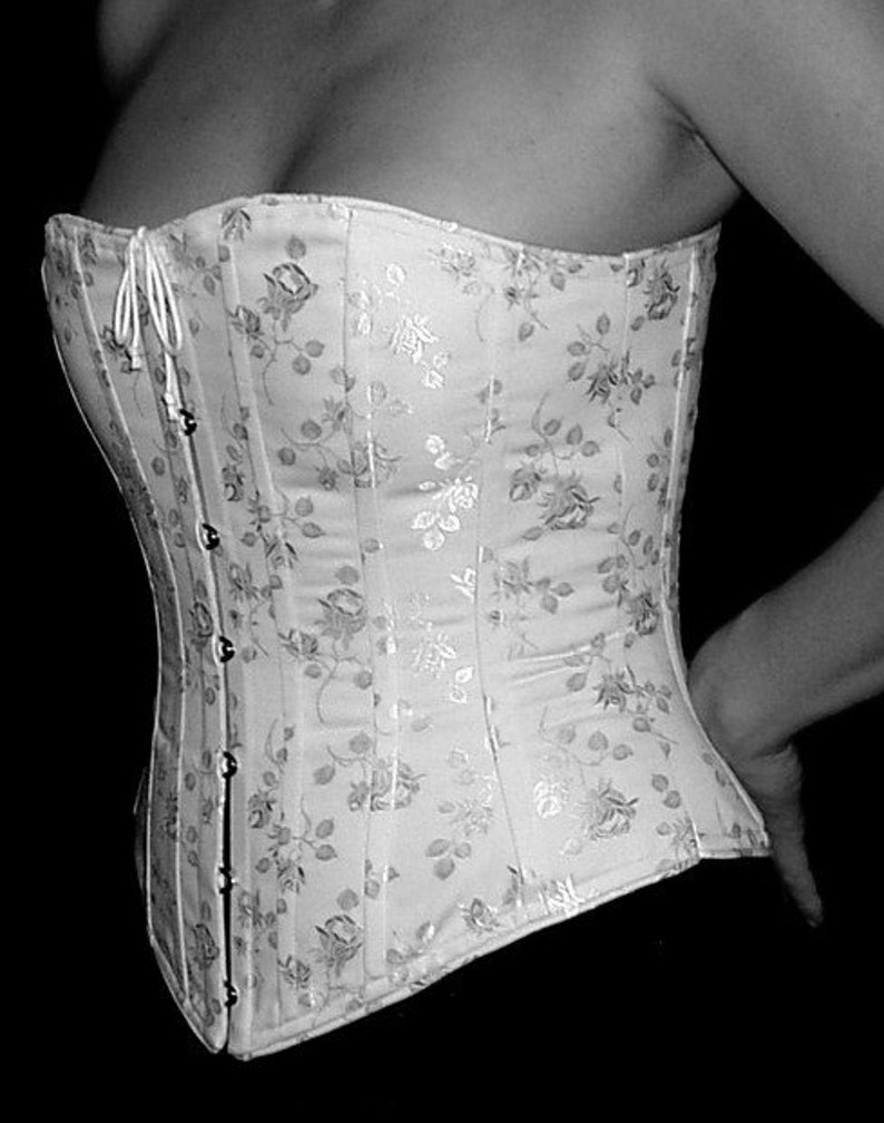 May include: A white corset featuring a tonal floral pattern with a subtle sheen. The garment includes vertical boning channels and a front closure with silver-toned metal busk clasps and a white cord lacing detail at the top neckline.