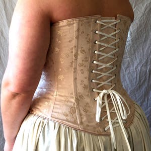 May include: A beige corset featuring a subtle floral jacquard pattern, constructed with vertical panels and silver-toned metal grommets. The back is secured with a white ribbon lace-up closure tied in a bow at the bottom, worn over a cream skirt.